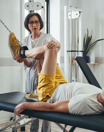 Mature man doing the knee flexion assisted by a qualified physical therapist at the gym
